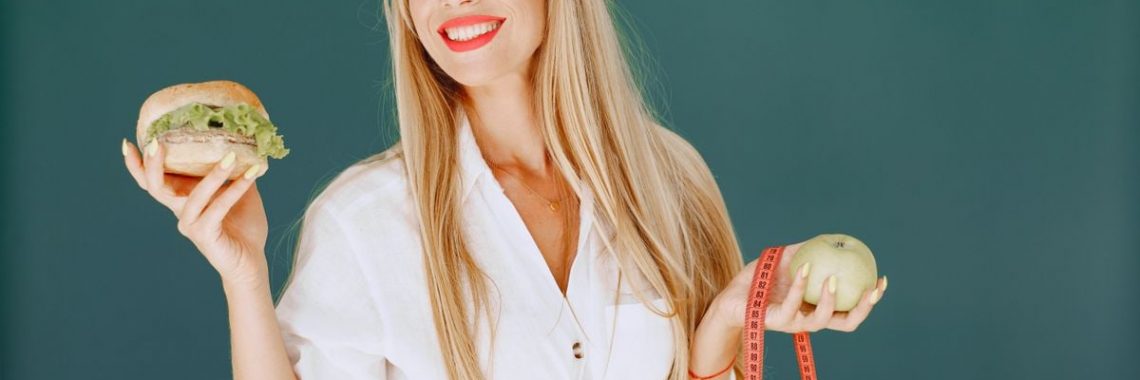 Smiling woman holding a burger and an apple, symbolizing healthy lifestyle choices.
